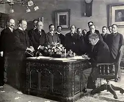  A black and white image of a group of men surrounding the Resolute desk, which had a large bouquet of flowers on it, as Jules Cambon signs the treaty on the desk.