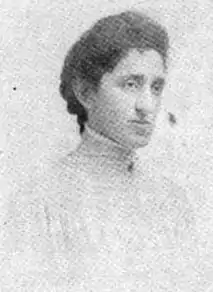 A woman with thick dark hair dressed back into braids at the nape; she is wearing a high-collared white lace shirtwaist or dress.