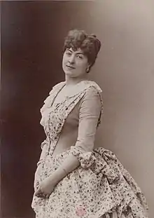 Sepia-toned portrait of a white woman, standing; her hair is dressed in curls atop her head; she is wearing a dress with calico-print skirt and front panel.