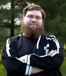 Justin Knapp—a Caucasian male with brown hair and a bushy beard—stands with his arms folded