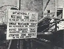 A multilingual sign (in German, Polish, Hungarian and French) warning people against trespassing for fear of being shot. Broken fence with some barbed wire appears behind the sign