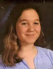 A smiling young preteen/teen girl with long, wavy brown hair and wearing a light blue striped shirt