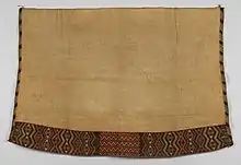 A photo of a woven cloak laid flag. It is mostly off-white, with geometric designs along its left, right, and bottom borders.