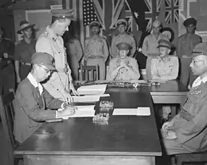 Men in uniform sit at a large wooden table. One is signing a document.