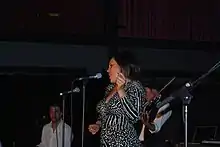 Upper body shot of a woman, singing and dancing before a microphone. She is shown in left profile and wearing a black and white dress. Behind her two male musicians are partly obscured or out of shot.