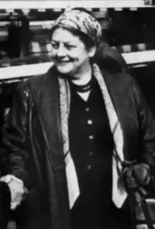 A middle-aged white woman, smiling and shaking someone's hand; she is wearing a print headscarf and a dark jacket over a darker dress