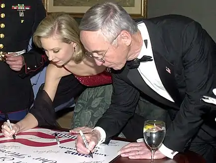 A young woman and an older man, both dressed in formal wear, leaning over to sign a poster.