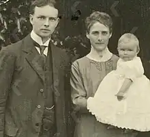 photograph of a man standing next to a woman, who is holding a baby