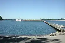 A small tugboat pushes a much larger barge around a safety marker at the end of a long row of pilings extending from a river bank. The tugboat is white with bits of red, while the barge is gray. Lettering on the side of the barge says "Tidewater".