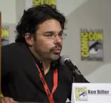 A man wearing a black/brown shirt speaks into a microphone while looking straight forward.