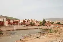 A river runs across the image from left to right with a town in the background, behind a concrete flood defence.  The foreground shows a stony, sparsely vegetated river bank.