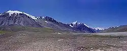 Some tall, blue-grey mountains rise out of brown soil below a deep blue sky