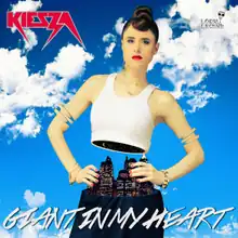 A portrait of a young woman with tied-up red hair portrayed as mannequin split in half from her waist area. Her backdrop is a blue sky smitten with white clouds. She sport a white tank top, navy pants and bright red lipstick. Below her in large white font stands the title in capital letters 'Giant in My Heart', and in the top-left corner of the portrait stands the woman's name 'Kiesza' in small bold red font, also in capital letters.