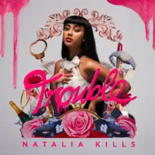 A young, tan-skinned woman wearing a white dress with her hand on her chest standing against a grey background. Around her are various items including  watches, champagne, police cars and roses. The word "Trouble" appears on the centre of the image. The text "Natalia Kills" appears at the foot of the image. Pink liquid flows down from the upper corners of the image.