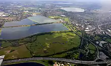 Seven reservoirs.  View of four in Spelthorne with small lakes of lower elevation, from aggregate extraction, in the south of the borough to the right.  Beyond three reservoirs in Elmbridge.  The flattest areas of the far north of the county. Staines road and rail bridges span the Thames into Runnymede in the right of the photograph.