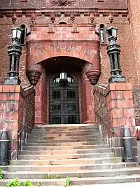 Stone steps, with high red stone block walls on either side and large black lanterns on those walls, leading up to a doorway with a black iron grille gate. Above it is a stone castle-like section on a brick facade, with the words "258th Field Artillery" in stylized lettering