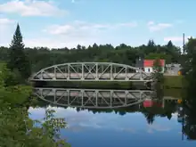 "A bridge crossing a river, the bridge reflects in the river. The steel girder bridge features an archway above the deck, with several beams interconnecting the arch with the deck.