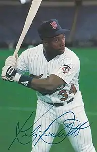 A man wearing a white baseball uniform stands holding a bat and ready to swing