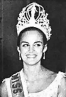 A smiling young woman with olive skin and dark hair, wearing a tiara and sash, and drop earrings