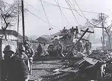 Soldiers moving through a damaged village with a tank following behind