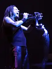 A man with his eyes closed and mouth open, holding a microphone; he is wearing dark clothing and wrist bands.