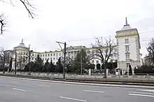 A white, four-floor palace is seen with side wings facing the viewer, each with a spire atop. A tram line and road can also be seen in front of the establishment.