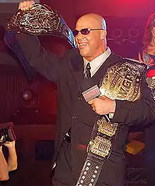 Kurt Angle with the original TNA World Heavyweight Championship title belt design with the IWGP Heavyweight Championship beside it.