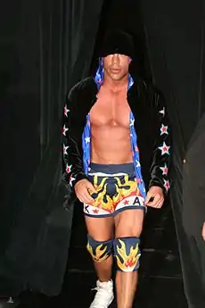 Adult white male wearing wrestling tights and knee pads as well as a black jacket.