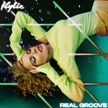 Kylie Minogue wearing a green dress, putting her hands on her head. Digital lights are edited in while her first name appears in the top left and the song title "Real Groove" appears in the bottom right.