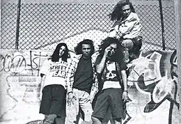 Four people, three of which are leaning against a wall and one of which is sitting on the wall. The wall has a lot of graffiti on it and there is a fence above the wall.