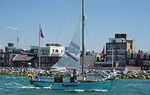 blue-hulled two-masted yacht with mainsail partly set passing a rocky shoreline with buildings beyond