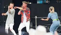 Members of L27, two white men, both wearing white pants, one wearing a red Hawaiian shirt and the other a gray t-shirt, sing into microphones on stage.