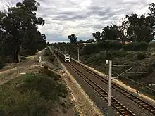 View from a bridge of a dual-tracked railway line with overhead power and foliage on either side indicating that construction had not begun yet.