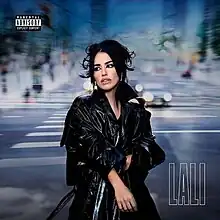 Lali wearing a black leather coat and her hair tied with a bun while looking over her shoulder and standing on a zebra crossing and blurred buildings behind her.