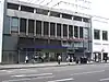 A grey building with blue panels and a dark blue, rectangular sign reading "LANCASTER GATE STATION" in white letters and people in front