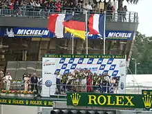 Above the race track, teams of three drivers are standing on the podium with national flags waving above them