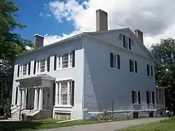 A pale blue house with black shutters, brown bric chimneys and a front porch with Greek-style columns seen from its front right.