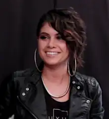 Still image of a woman looking away from the camera and smiling with an undercut hairstyle; wearing large hoop earrings, a black top, a thick necklace, and a leather jacket.