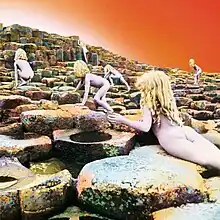 Six nude children with long blonde hair scramble up a stairstep series of basalt rocks ascending away from the viewer, with an orange-white sky above