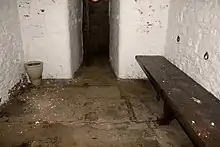 A police cell, with white walls, stone floor and a bench