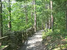  A gravel path through a mixed forest of deciduous and conifer trees, with a rail fence supported by stone pillars left of the path