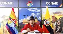 A man in a fedora and poncho, seated in front of logos for CONAIE and the flags of CONAIE and Ecuador