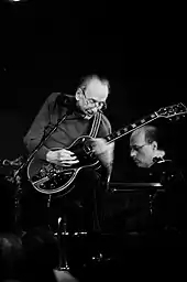 Black and white image of a man behind a microphone stand holding a guitar; in the background is a man at a piano