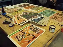 Colorful letterpress posters for "St Louis Microfest" spread out on a table alongside ink and other printing materials.