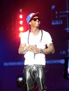 A smiling black male wearing sunglasses and a baseball cap stands in front of several stage lights.