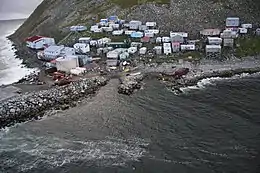 A village of 40–50 mostly small buildings built on a steep, rocky coastline; some structures higher on the hill are built on stilts.