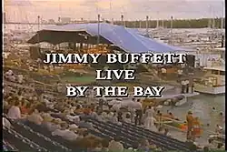 A screenshot of the opening titles of Live by the Bay; in the foreground are partly filled bleachers (stands); the center of the image is the canvas-covered stage of Miami Marine Stadium; in the water surrounding the stage are numerous pleasure boats; the skyline of Miami is barely visible in the top left of the frame; the text "Jimmy Buffett Live by the Bay" appears capitalized in plain text superimposed on the frame