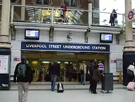 A white building with a dark blue, rectangular sign reading "LIVERPOOL STREET UNDERGROUND STATION" in white letters all under a white sky