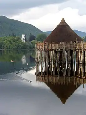 A small, brown conical structure sits on top of wooden piers set into a body of water. Ducks paddle through the water and in the near background there is a tree-lined shore with a white square tower showing amongst the trees. Tree covered hills and grey skies dominate the far background.