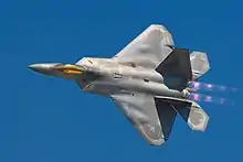 Dorsal view of F-22 in a steep bank, ailerons clearly banking wings, modern camouflage, bright gold canopy, jet thrust possibly purple with afterburner and cloud-effect from leading edges of wings all suggesting high speed